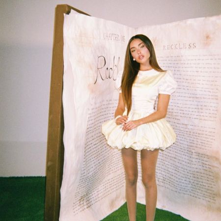 Madison Beer posing solo in a white dress in front of a giant prop. 